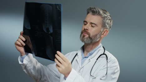 Caucasian-gray-haired-good-looking-man-doctor-holding-X-ray-copy-and-studying-it-carefully-on-the-grey-background.